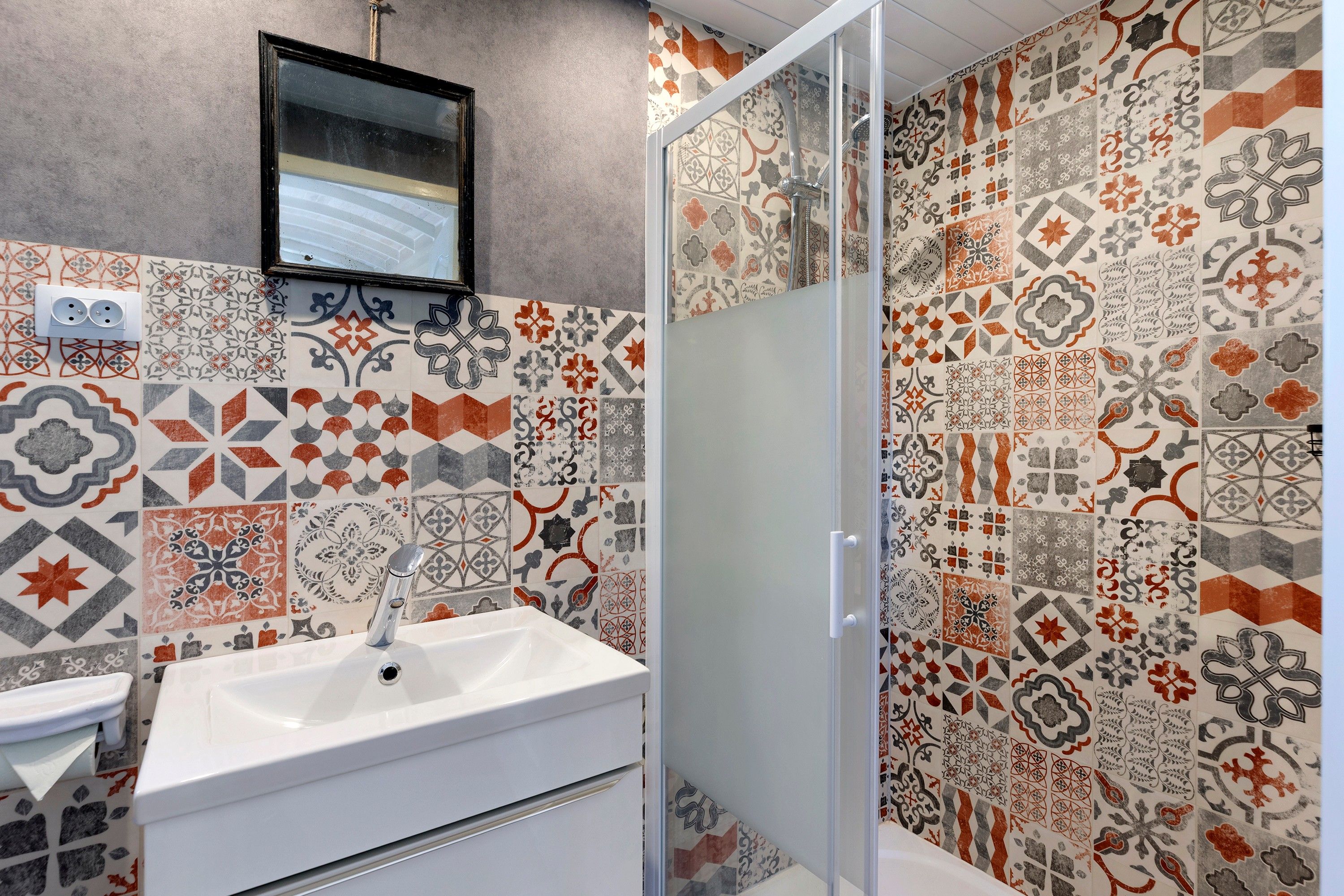 Une salle de bain avec un lavabo blanc, un miroir et une douche dotée d’une porte en verre. Les murs sont recouverts de carreaux à motifs de différentes couleurs.