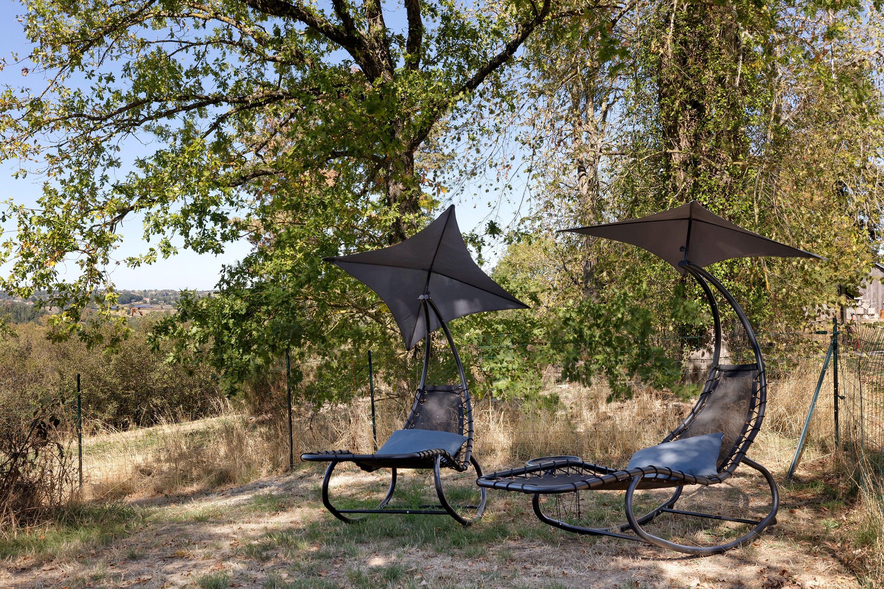 Twee buitsschommels met parasols, voor een achtergrond van bomen en een heldere lucht.