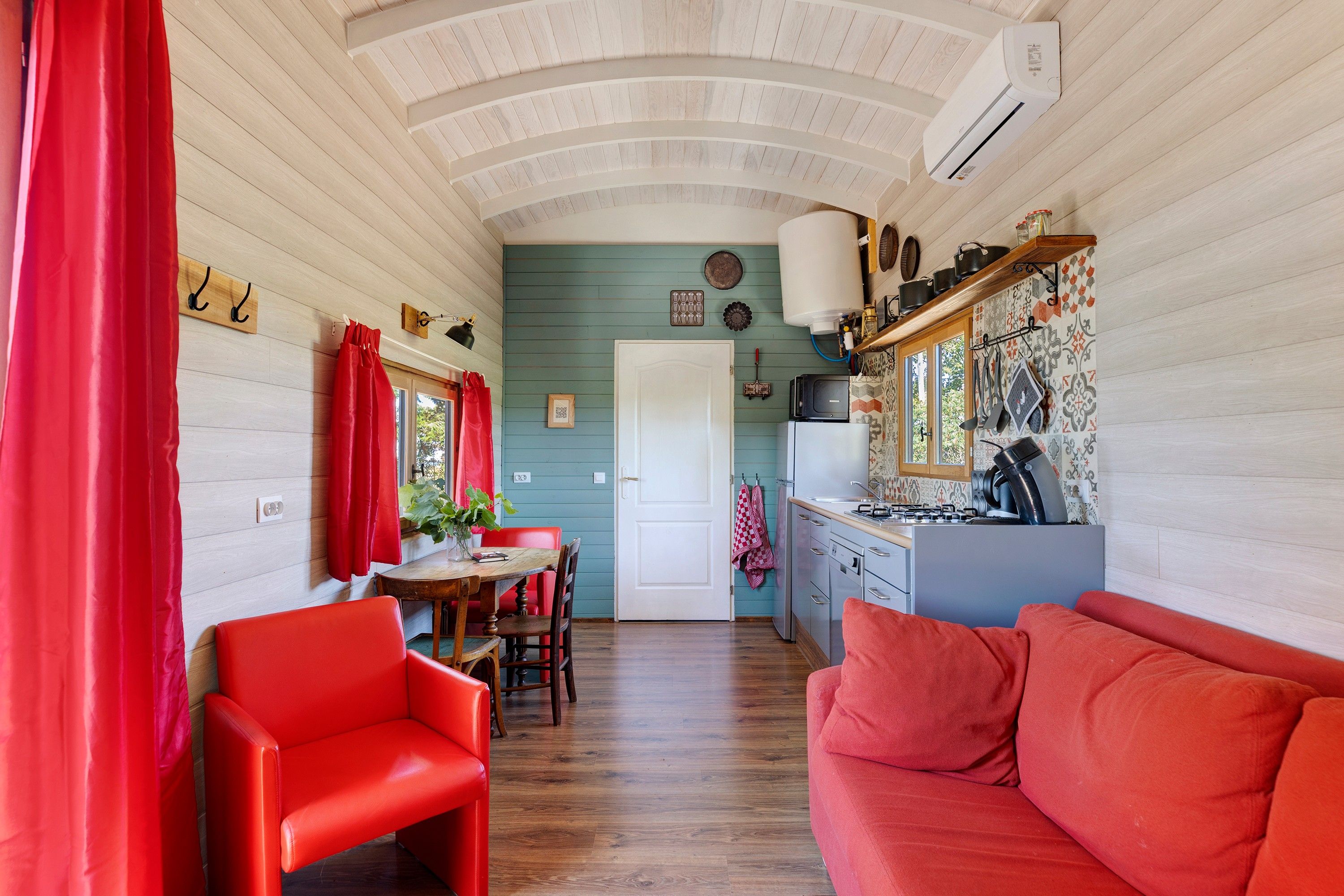 A compact kitchen with a dining area, featuring a wooden ceiling, a white door, and a red sofa.