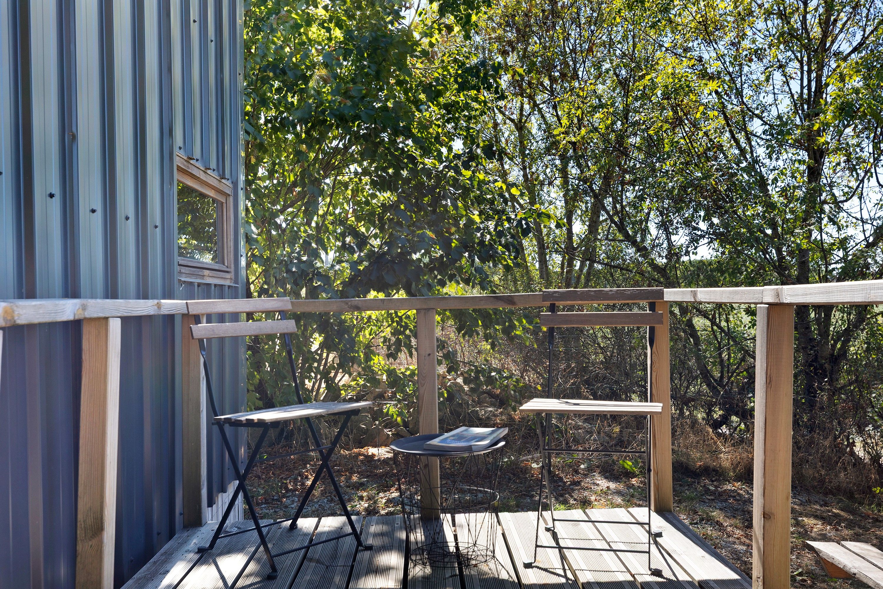 Une terrasse en bois avec une table et des chaises en métal, entourée d’arbres.