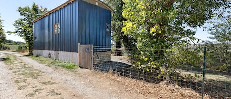 A blue metal building with a wooden door and a small window, surrounded by trees and a gravel path.