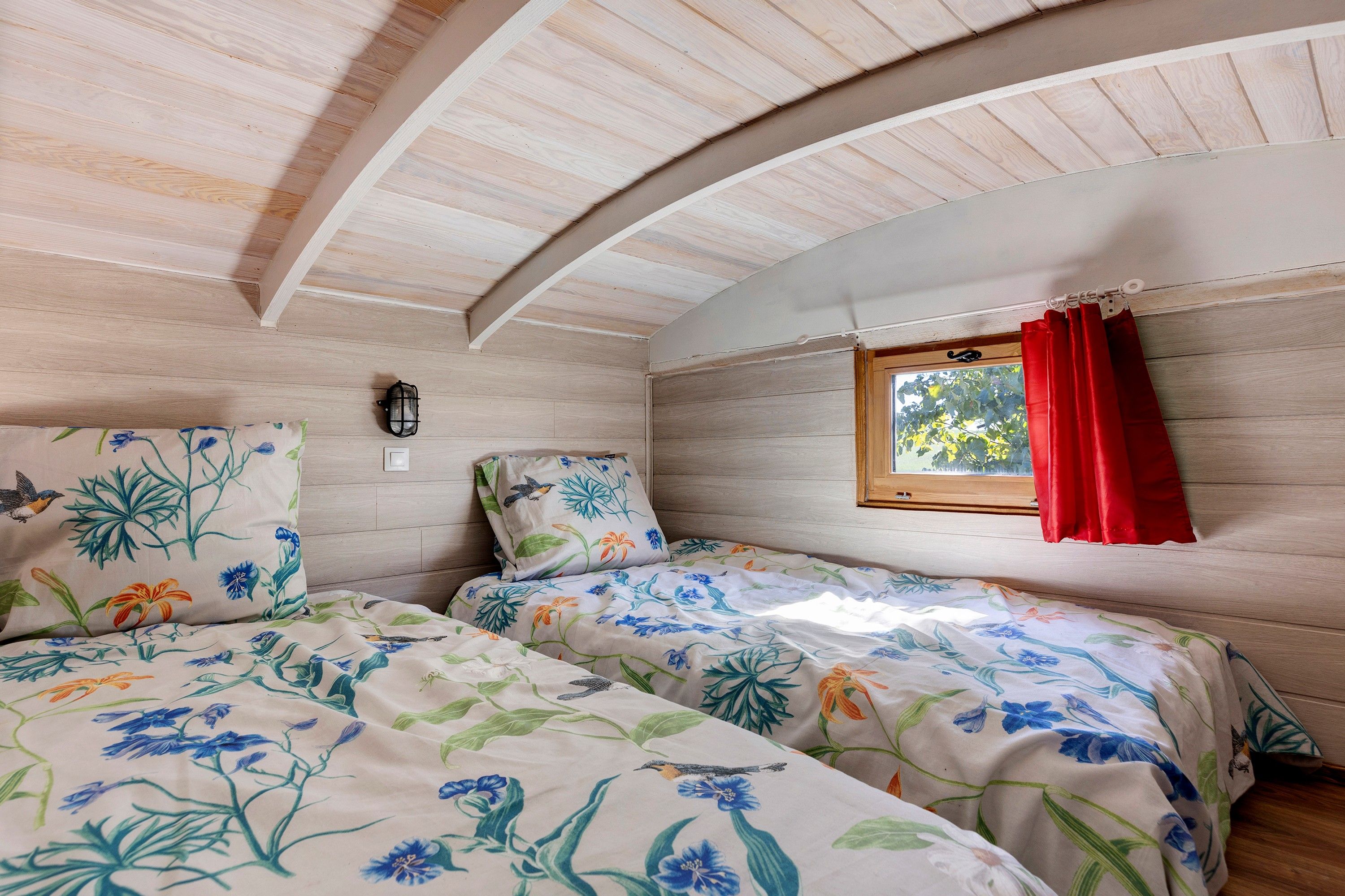 Un dormitorio con dos camas, ropa de cama floral, una ventana con cortinas rojas y una lámpara fijada a la pared.