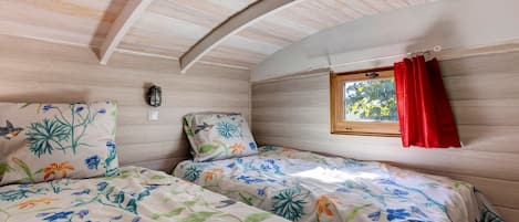A bedroom with two beds, floral bedding, a window with red curtains, and a wall-mounted light fixture.