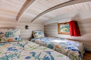 A bedroom with two beds, floral bedding, a window with red curtains, and a wall-mounted light fixture. - Luxury Caravan in the Heart of Nature (Charron)