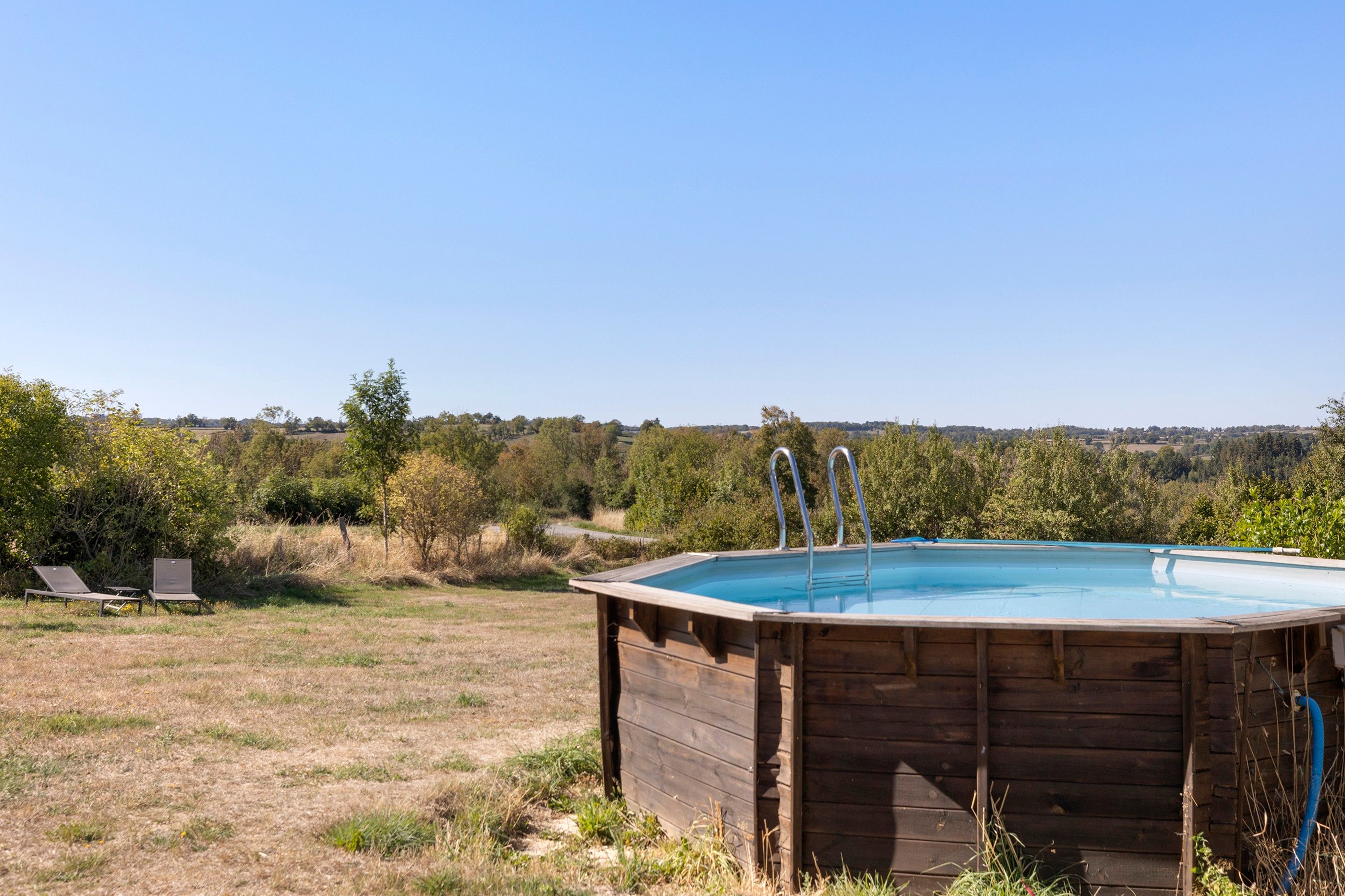 Ein hölzerner Pool mit zwei Leitern, umgeben von einer grünen Wiese und Bäumen, mit blauem Himmel darüber.