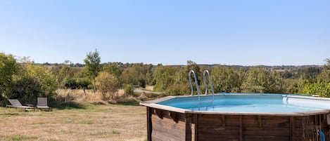 A wooden swimming pool with two ladders, surrounded by a grassy field and trees, with a clear sky above.