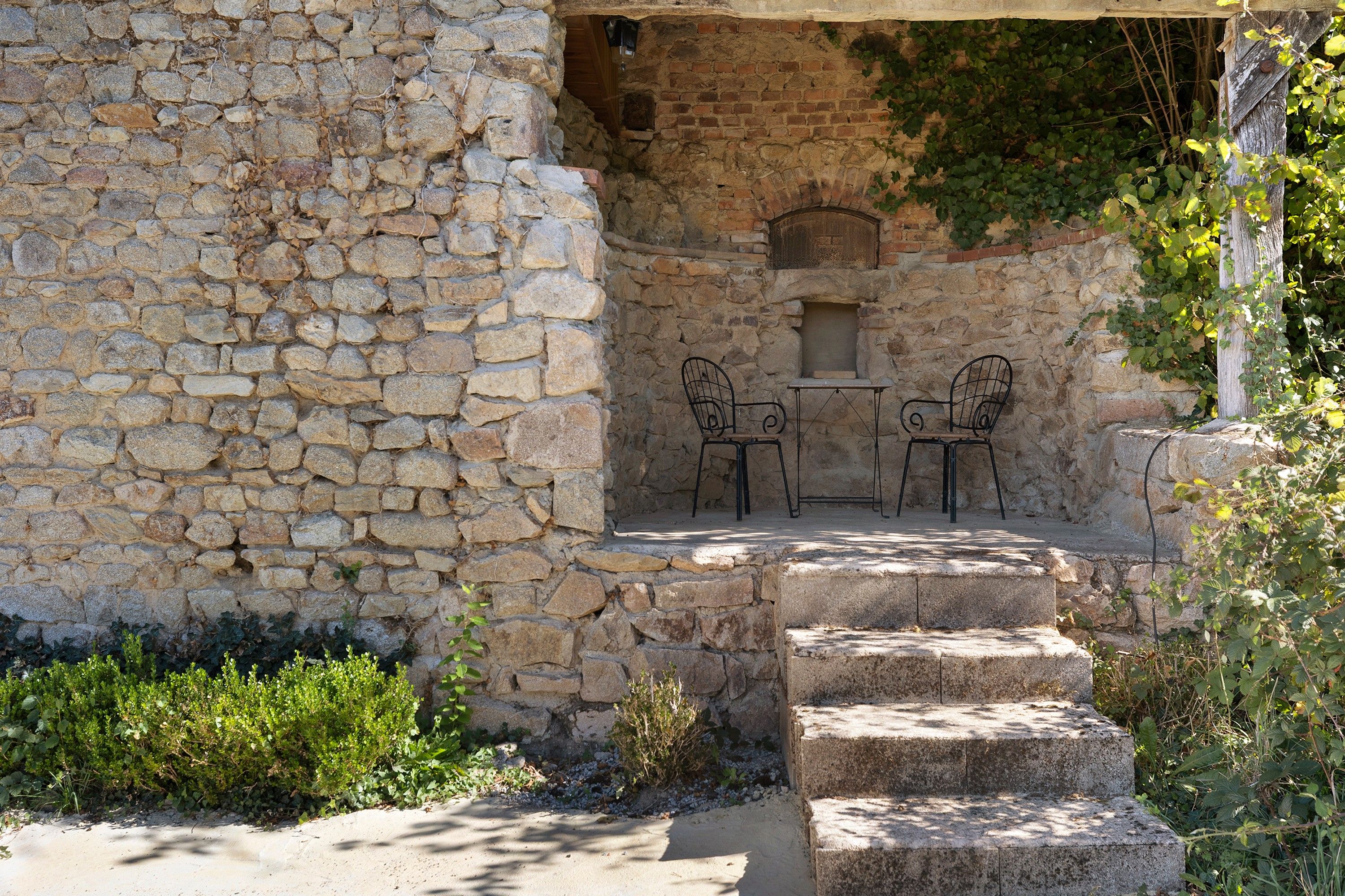 A stone building with a stone staircase leading to a small patio area with two metal chairs and a table.