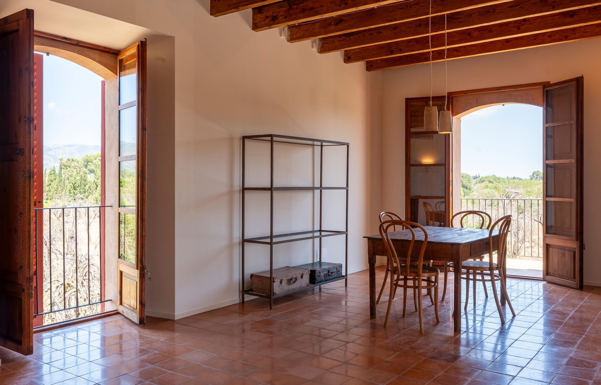Un coin repas avec une table et des chaises en bois, une étagère en métal et une vue sur l’extérieur grâce à une porte ouverte.