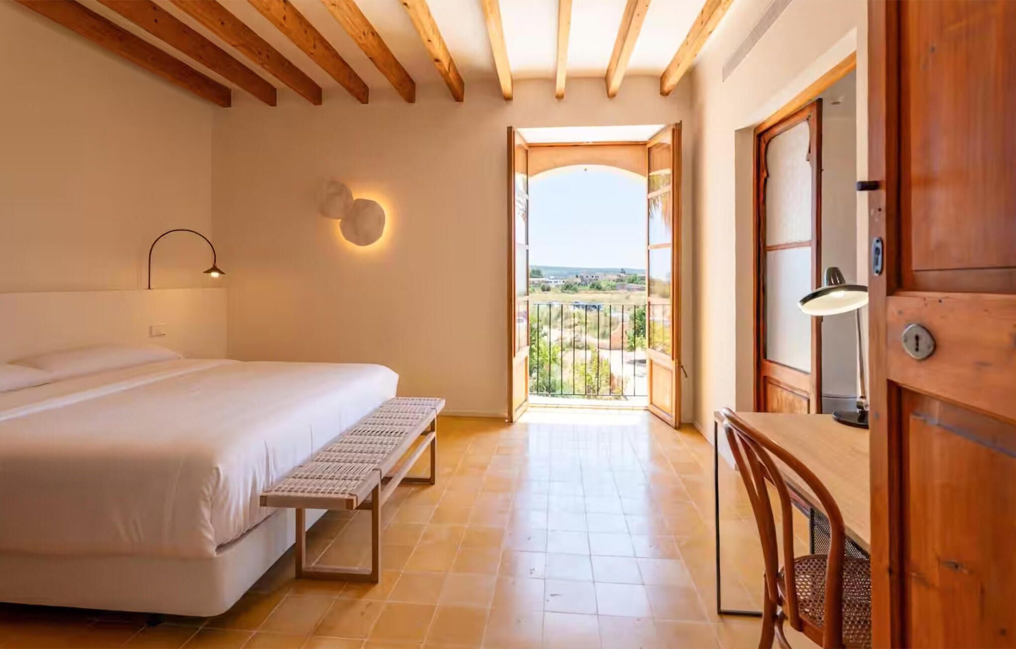 A bedroom with a bed, a bench, a desk, and a door leading to a balcony with a view.
