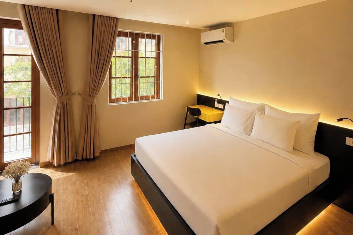 Quarto de hotel com uma cama grande, uma escrivaninha e uma cadeira. Há uma janela com cortinas e um ar condicionado na parede.