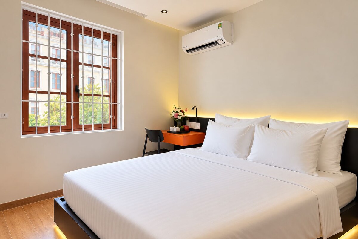 Een hotelkamer met een groot bed, een bureau met stoel, een nachtkastje en een raam met uitzicht op gebouwen en groen.