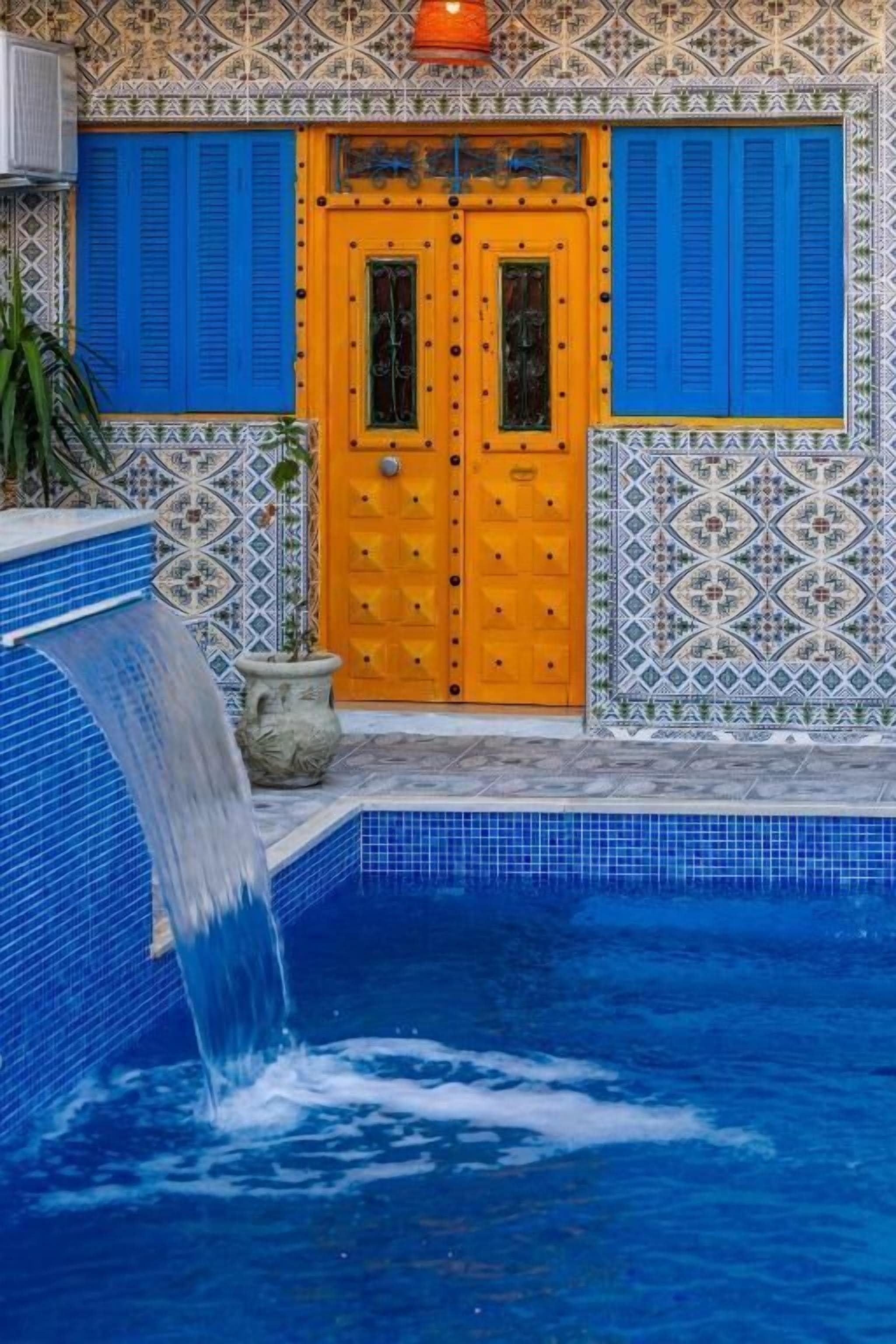Una piscina con cascada, una puerta de madera con contraventanas azules y paredes de azulejos decorativos.