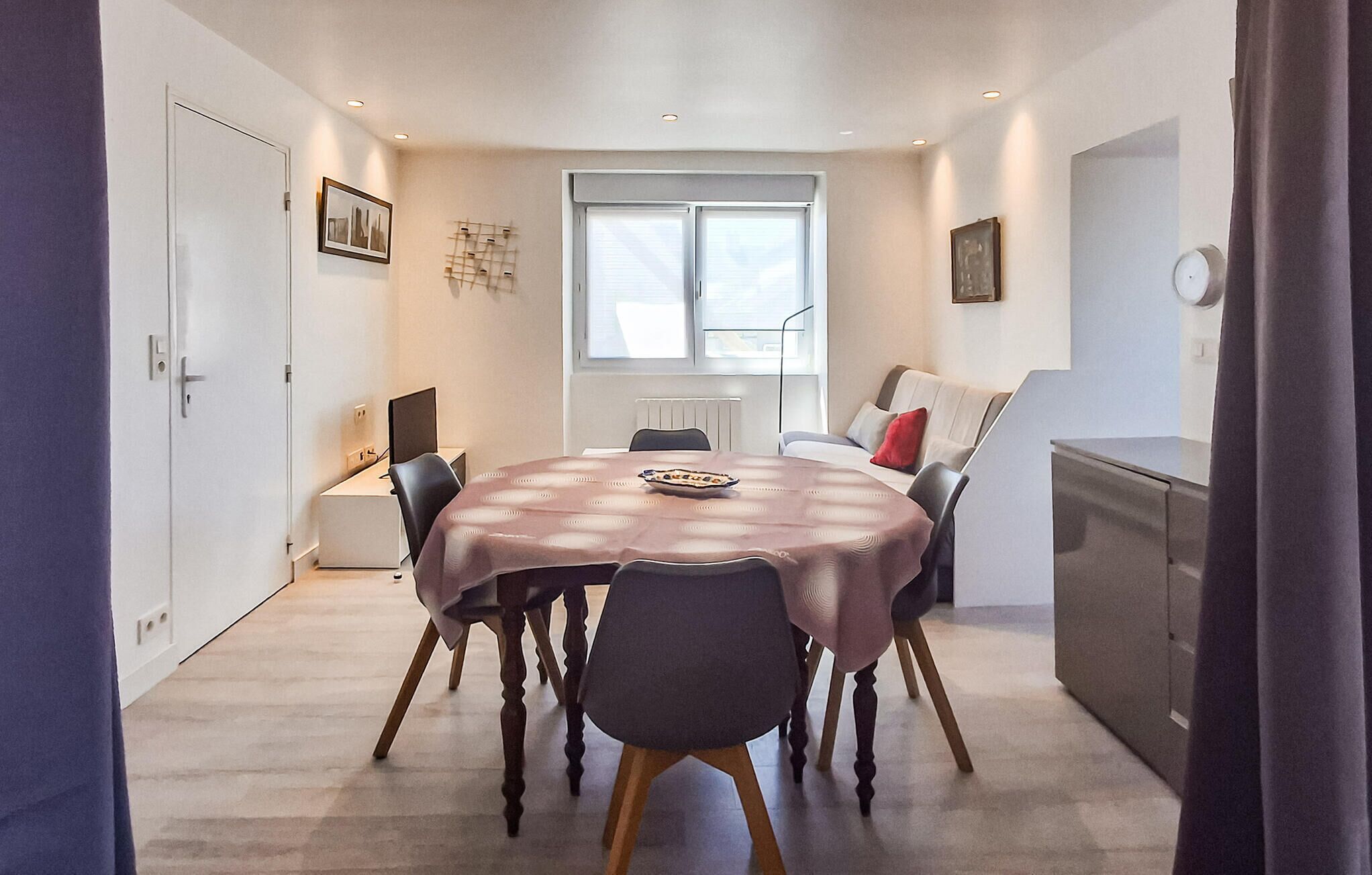 A dining area with a round table, chairs, a window, and a kitchenette.