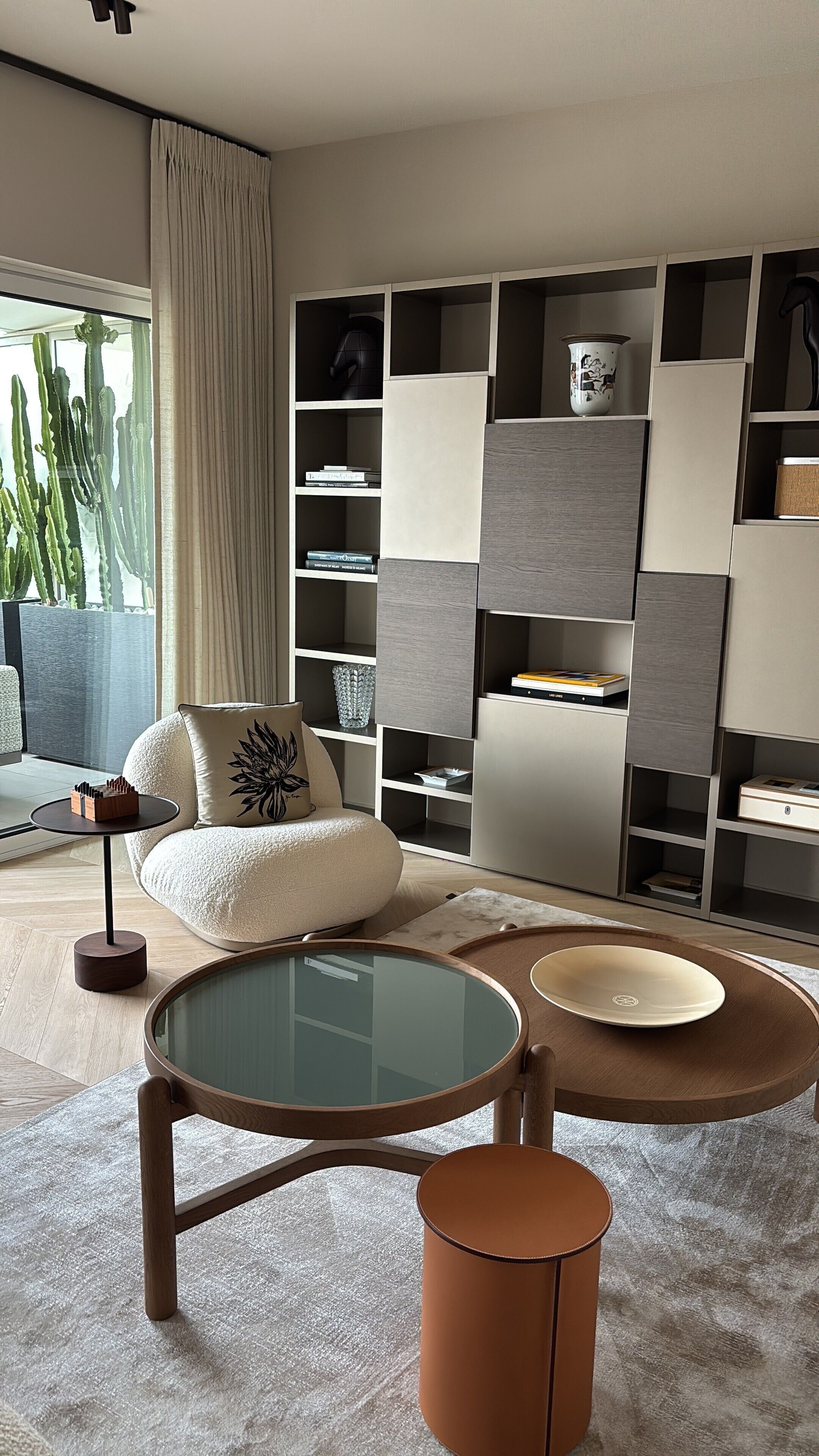 A modern living room with a sofa, a glass-top coffee table, and a side table. There is a bookshelf with various items and a large window with a view of cacti.