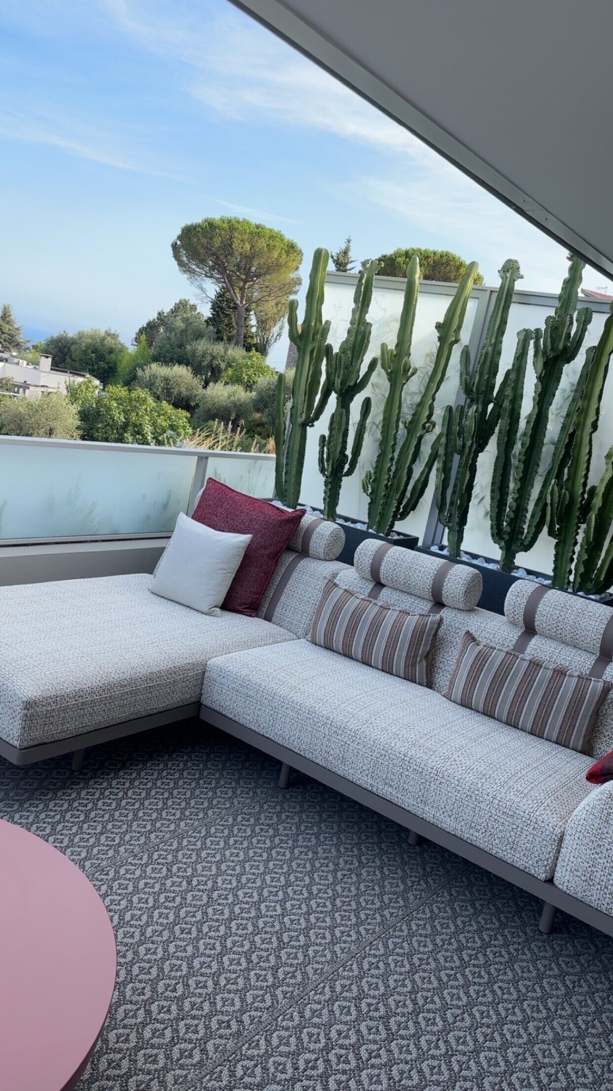 A modern outdoor seating area with a sofa and a round table, overlooking a garden with cacti and trees.