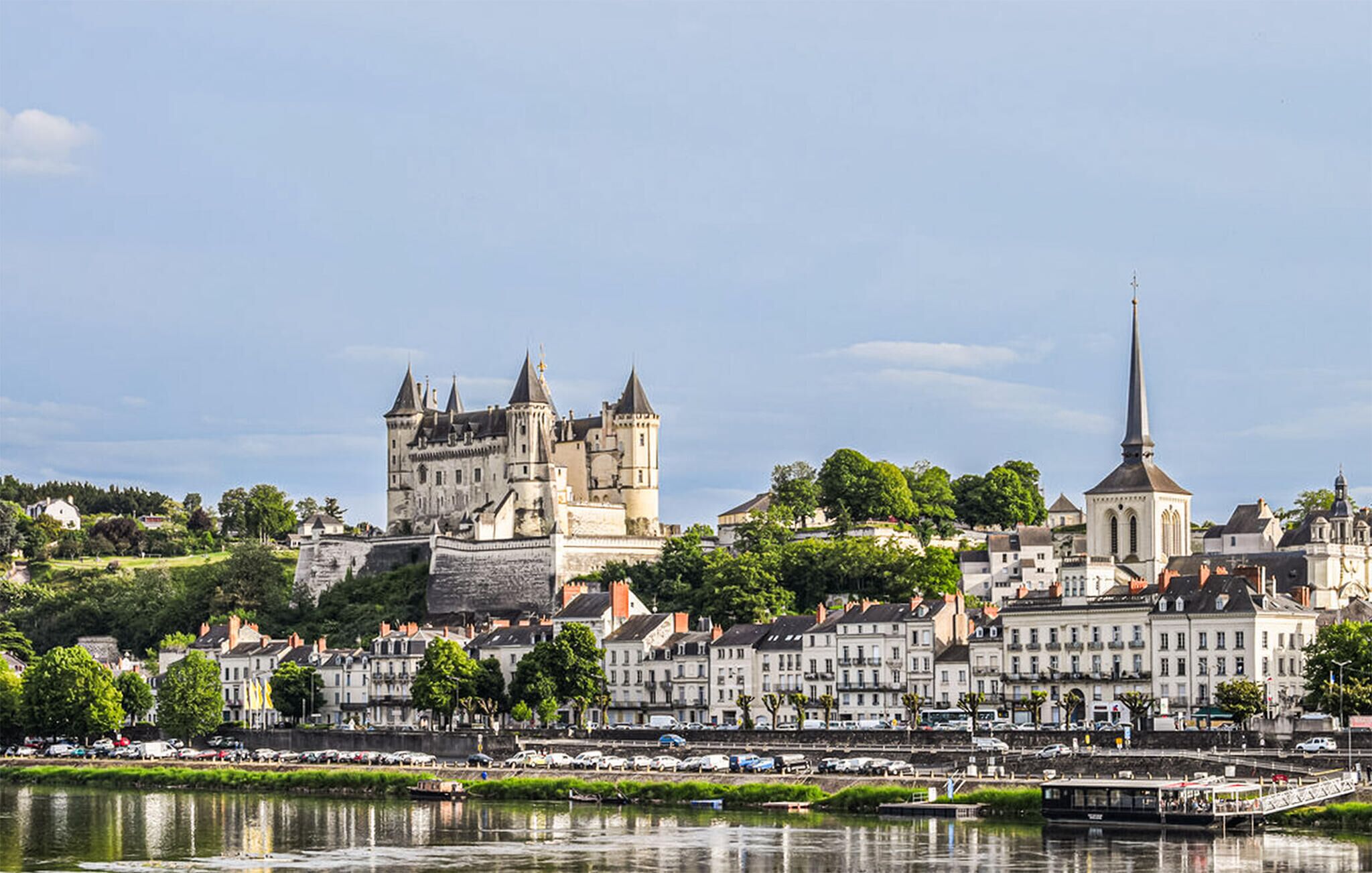 Eine historische Burg und eine Kirche mit hohem Turm, umgeben von einem Fluss und einer Stadt mit Häusern am Flussufer.