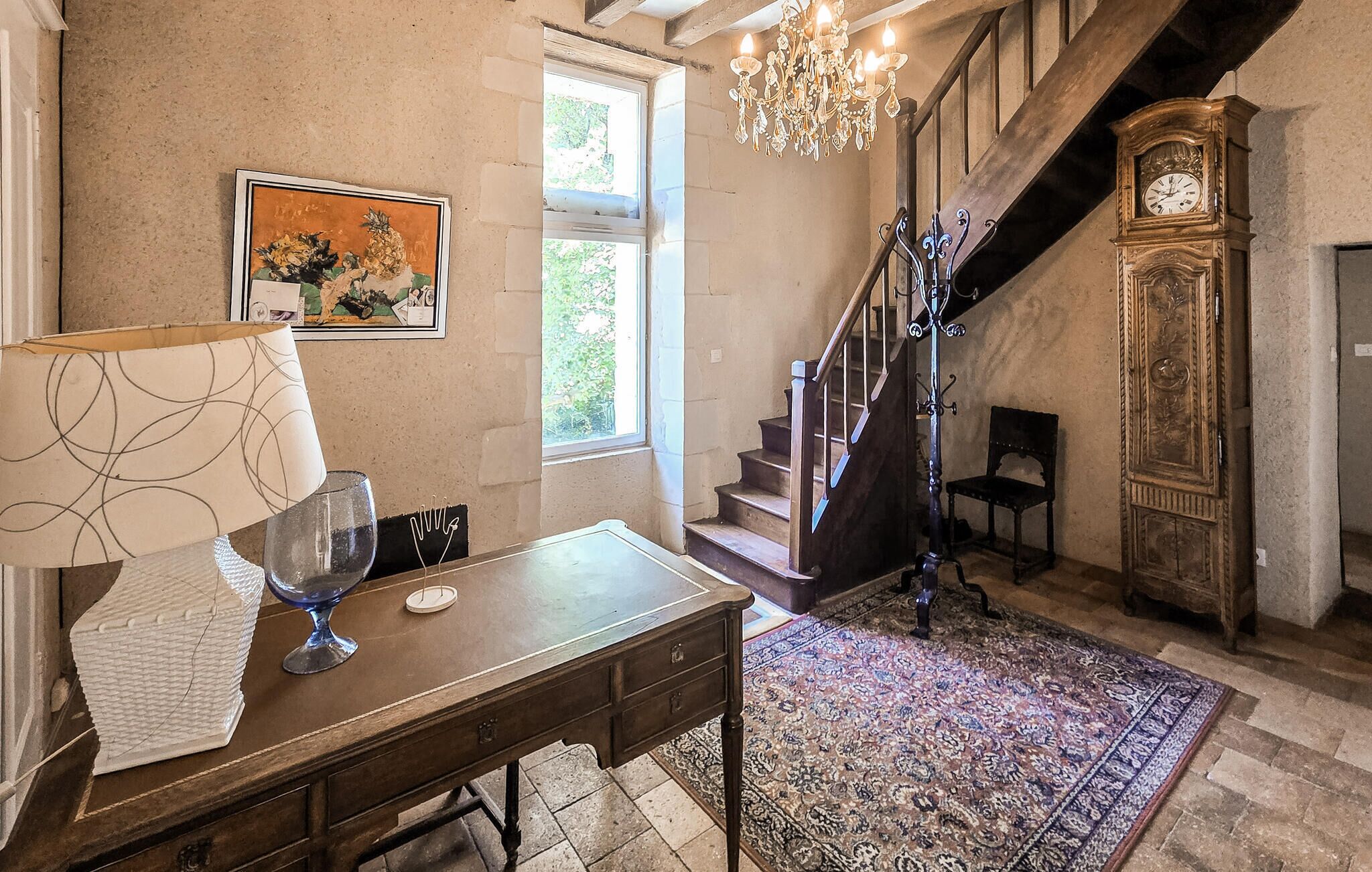 Ein Zimmer mit einem Holztisch, einer Kristallleuchter, einer Standuhr und einer Treppe.