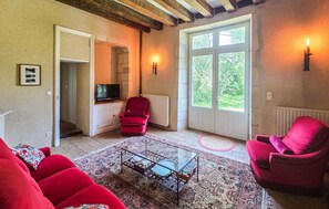 Ein Wohnzimmer mit Fernseher, roten Sesseln, einem Glastisch und einem großen Fenster mit Blick auf Grünflächen.