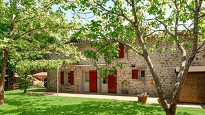Un bâtiment en pierre avec des volets rouges, une pelouse et un arbre devant.