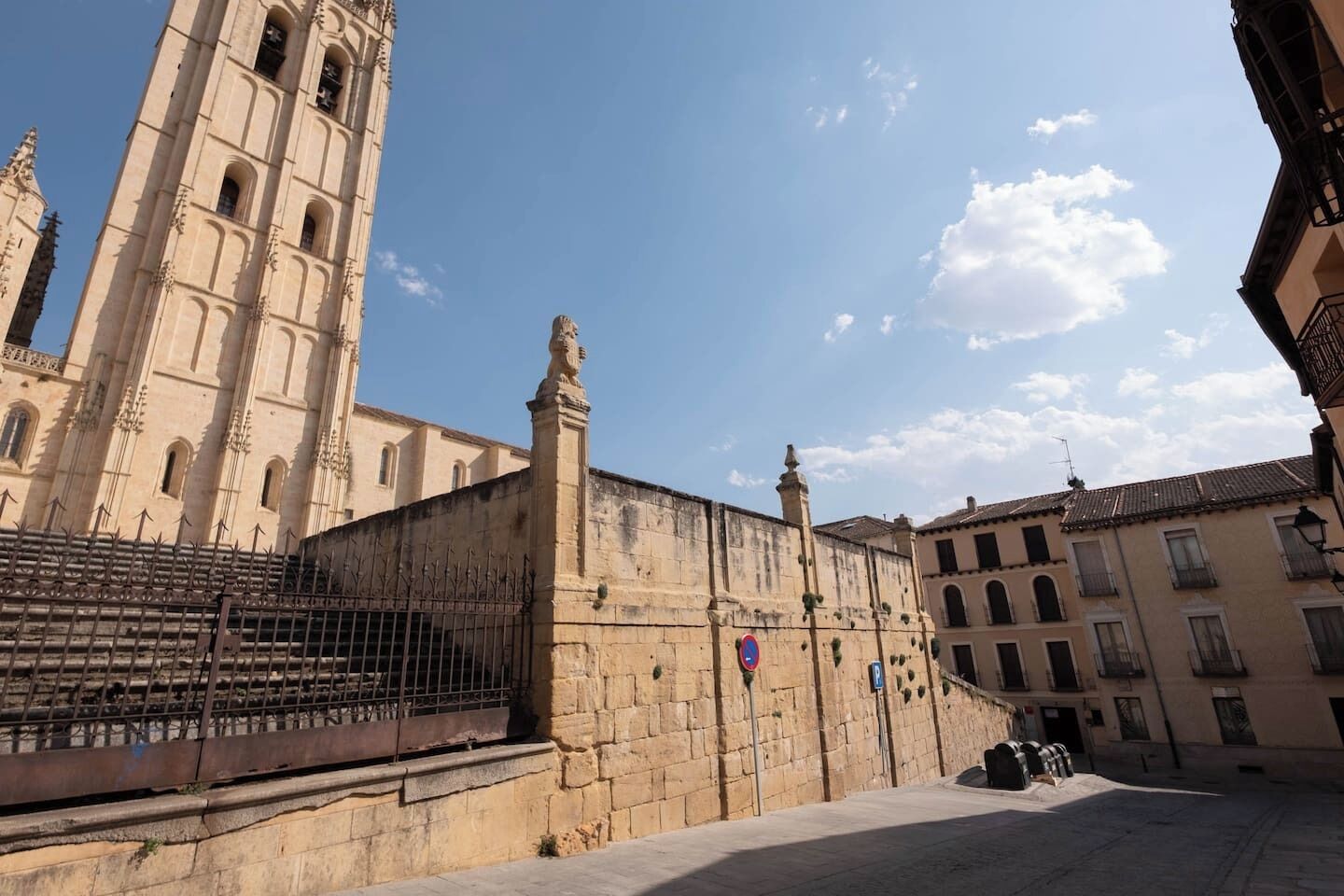 Un edificio histórico con un alto campanario, una muralla de piedra con una puerta y una plaza pavimentada.