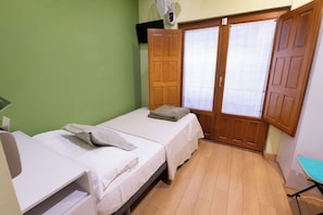 A small room with two beds, a wooden wardrobe, a window with blinds, and a small table.