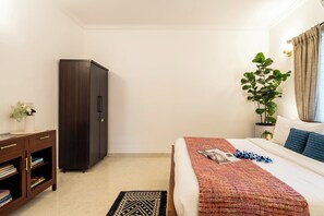 A bedroom with a bed, a wardrobe, a bedside table with a lamp, a potted plant, and a window with curtains.