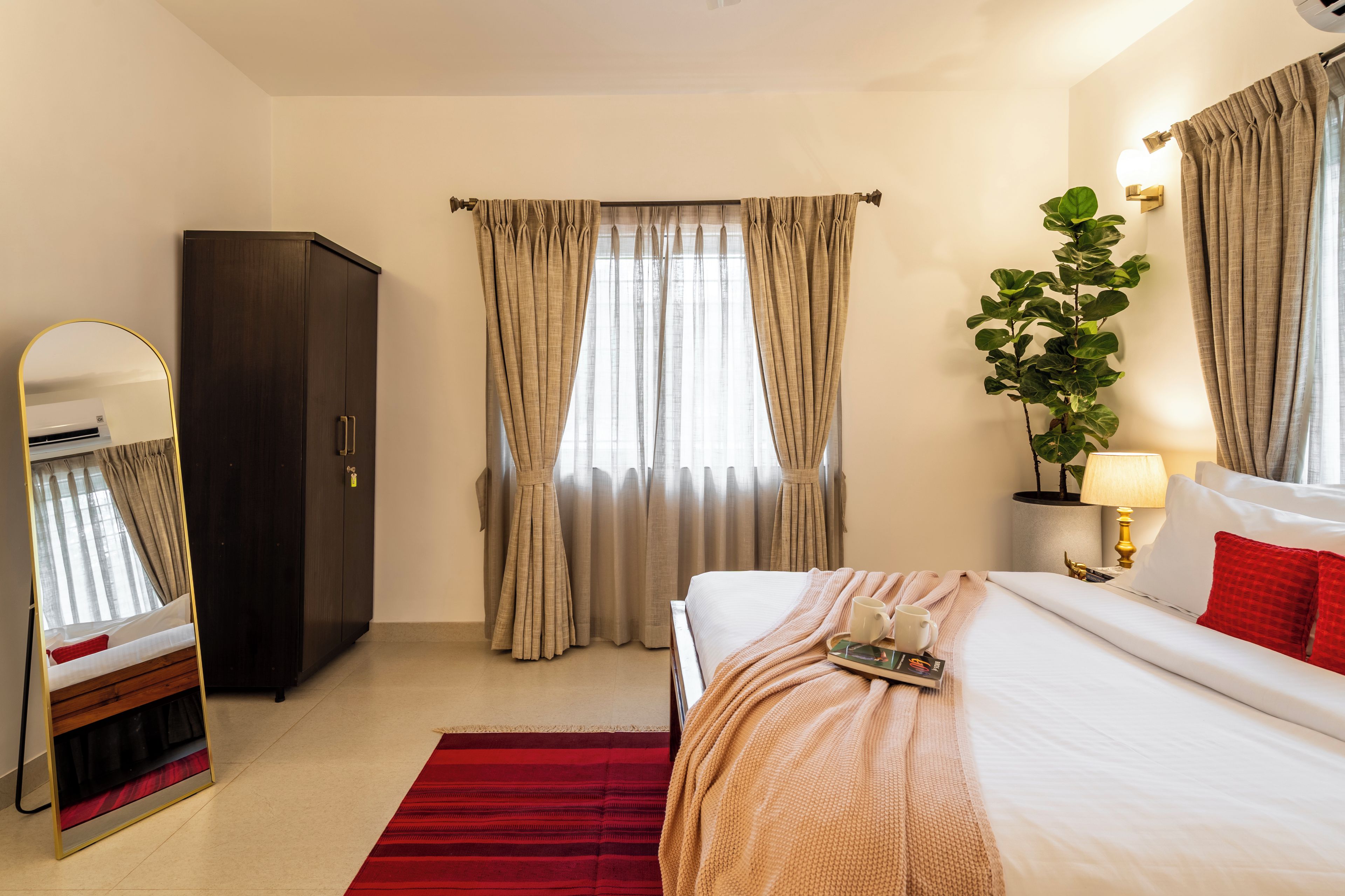 Ein Hotelzimmer mit Bett, Schrank, Spiegel, einem Fenster mit Vorhängen, einer Pflanze und einem Nachttisch mit Lampe.
