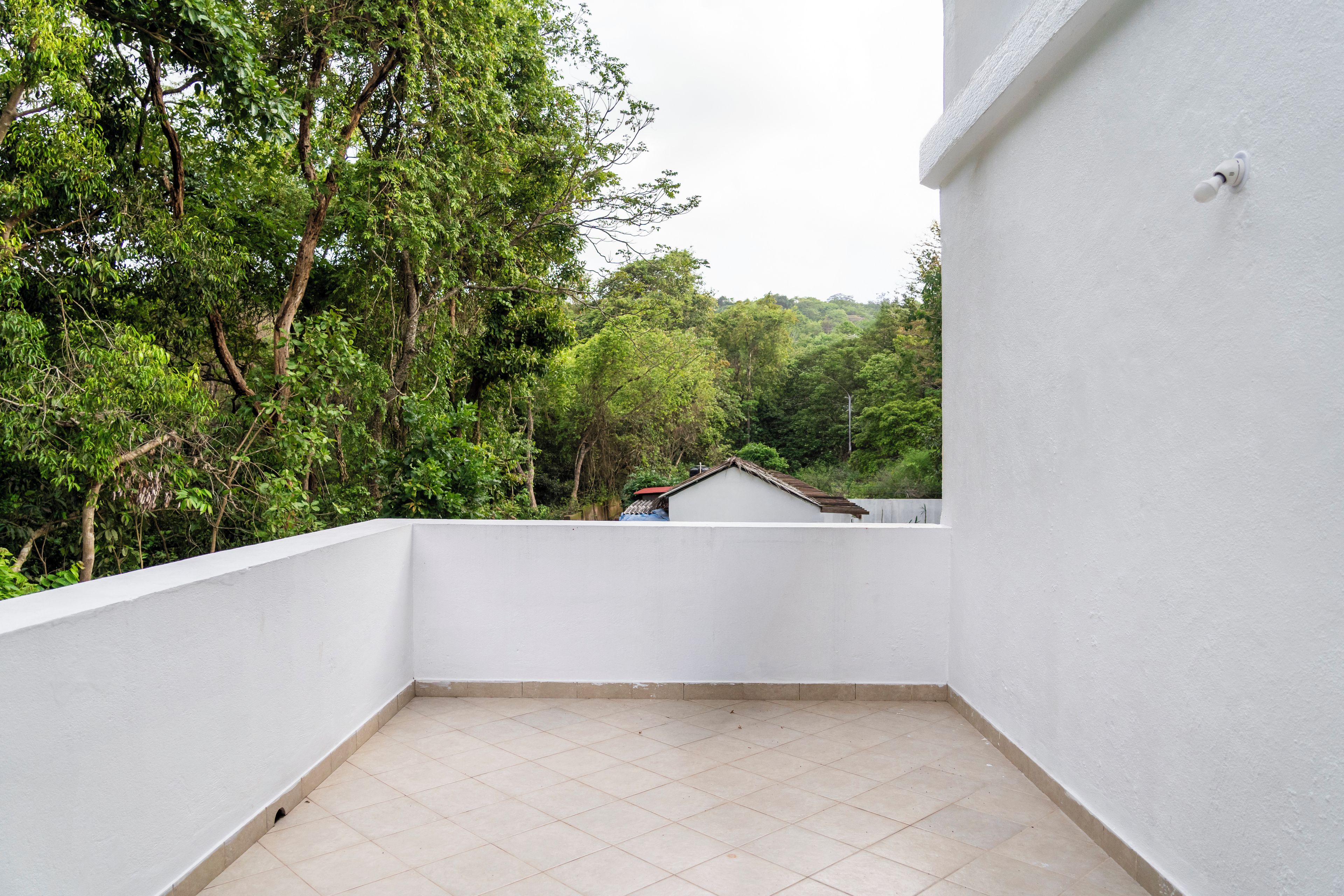 A balcony with a tiled floor, surrounded by white walls and a view of dense green foliage and a distant building.