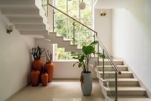 A staircase with a glass railing, potted plants, and a large window with a view of greenery. - Staymaster Caetano-3BR Villa with Pool  (Siolim)