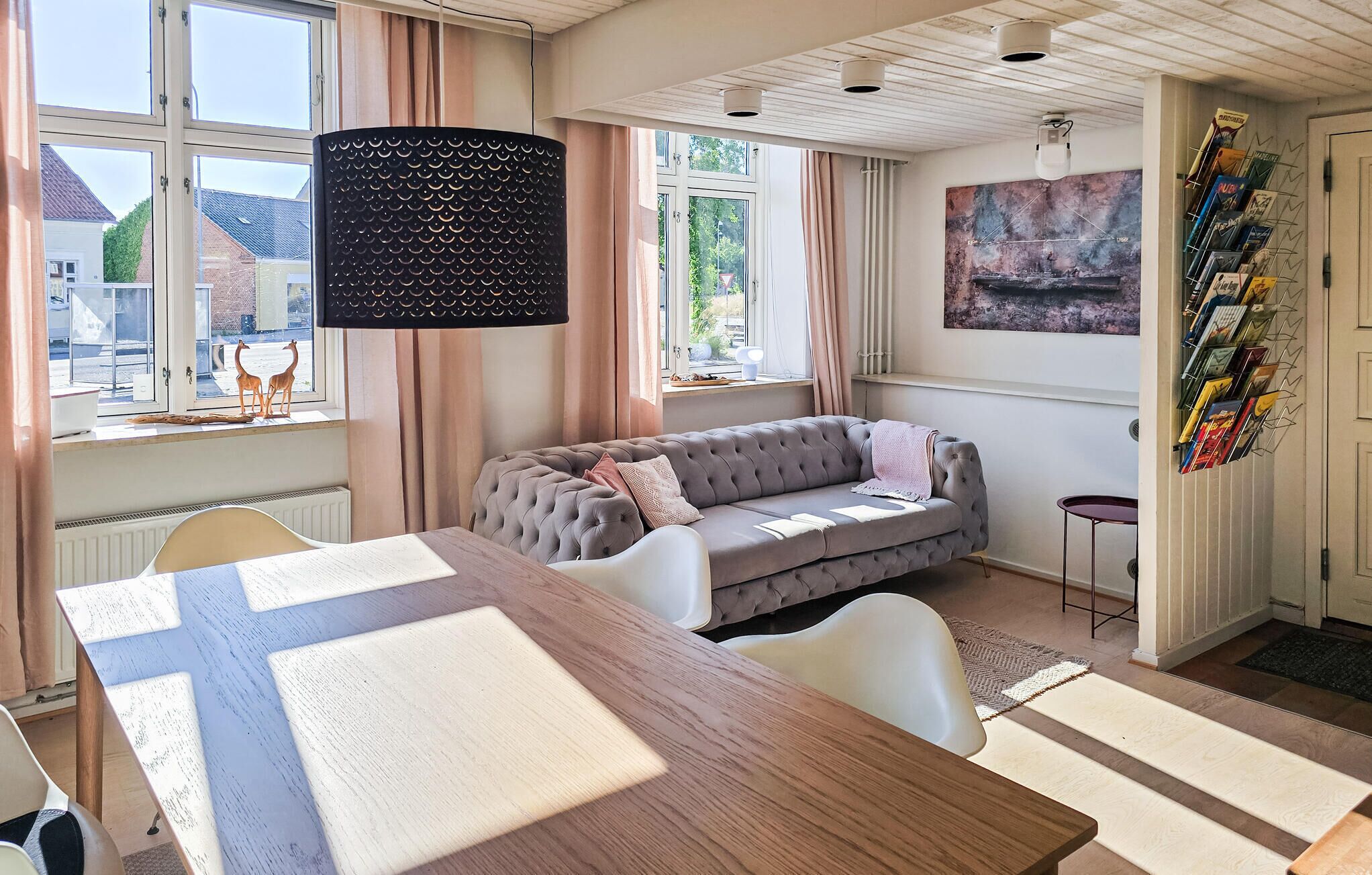 Un saló modern amb un sofà gris, una taula de dinar de fusta i un gran finestral amb cortines.
