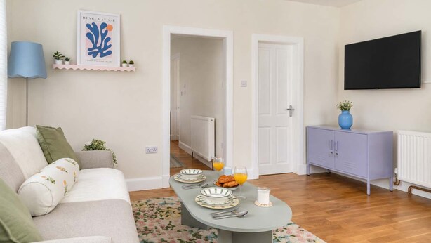Ein Wohnzimmer mit einer Couch, einem Esstisch gedeckt mit Tellern und Gläsern, einer blauen Lampe, einem gerahmten Kunstwerk und einem an der Wand befestigten Fernseher.