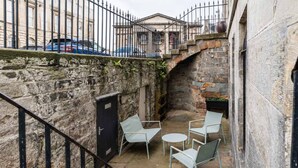 A stone-walled courtyard with a metal staircase, a black door, and outdoor seating.