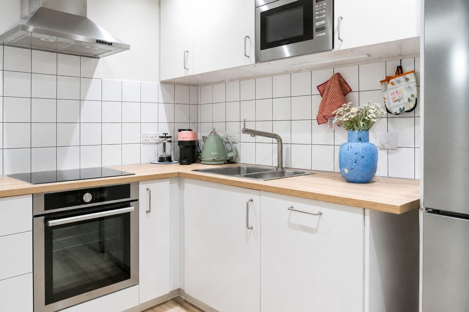 Moderni keittiö, jossa on valkoiset kaapit, ruostumattomasta teräksestä valmistetut kodinkoneet ja puinen työtaso.