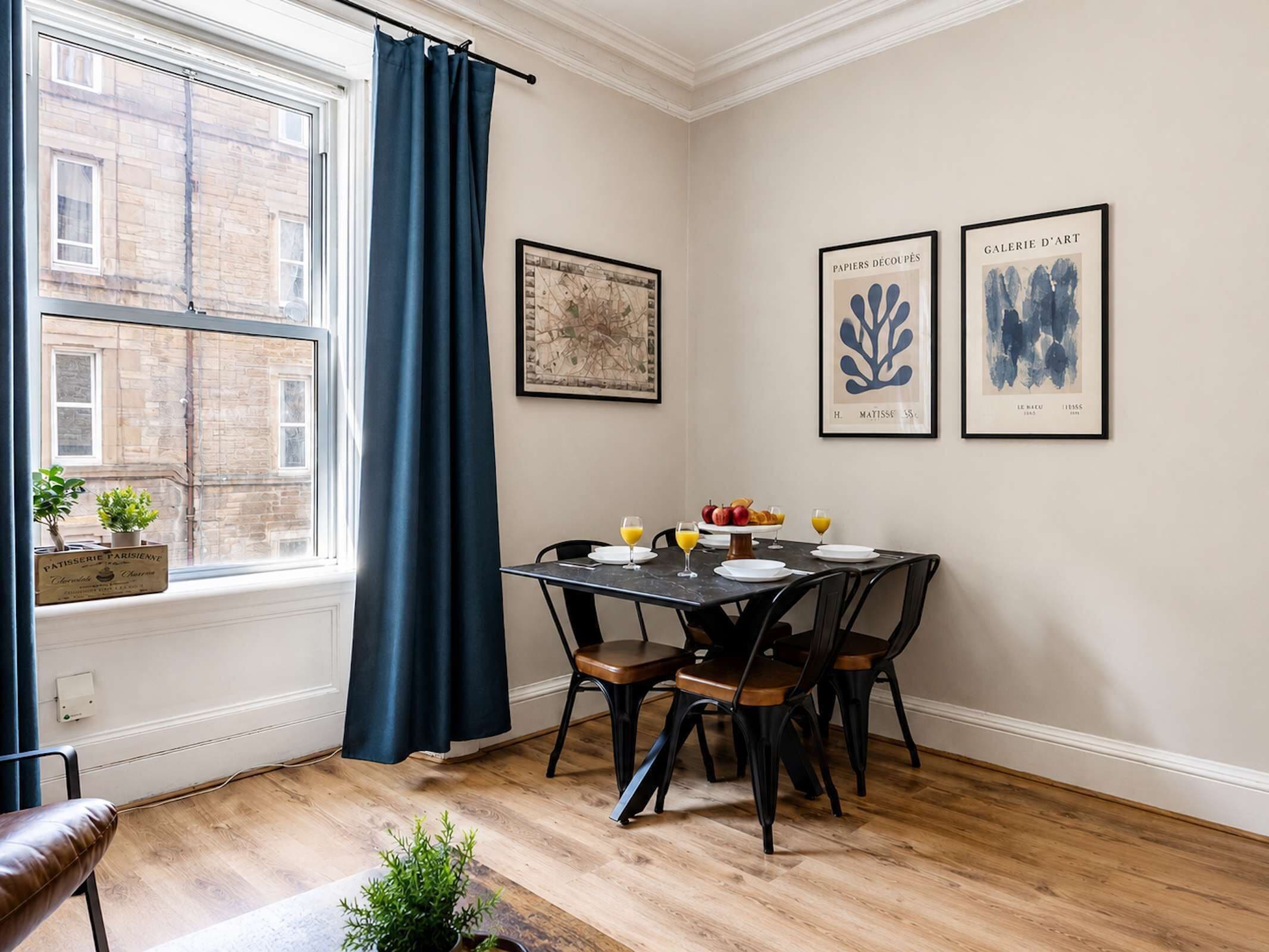 A dining area with a table set for two, a window with curtains, and artwork on the wall.