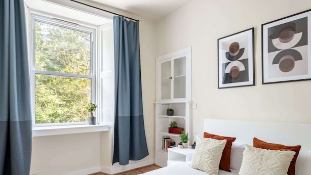 A bedroom with a bed, a nightstand, a window with curtains, a bookshelf, and two framed pictures on the wall. - Stylish 2BR Walk to Edinburgh Castle (Edinburgh)