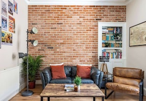 A living room with a brick wall, a leather sofa, a coffee table with books, a bookshelf, and a potted plant. - Stylish 2BR Walk to Edinburgh Castle (Edinburgh)