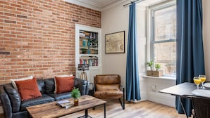 A living room with a brick wall, a leather sofa, a coffee table, a bookshelf, a chair, a window with curtains, and a small table with glasses. - Stylish 2BR Walk to Edinburgh Castle (Edinburgh)