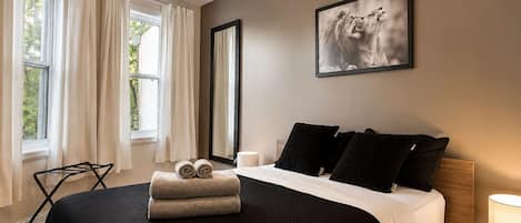 A bedroom with a bed, a large window, a painting, and a nightstand with a lamp.