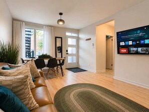 Una sala de estar moderna con un sofá, un comedor y un televisor mostrando la interfaz de un servicio de streaming.