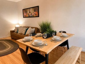 Un comedor con una mesa de madera dispuesta para cuatro personas, rodeada por sillas negras, un sofá marrón y un cuadro sobre la mesa.