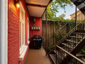 Una terraza de madera estrecha con un muro de ladrillo rojo, una parrilla negra y una escalera de metal.
