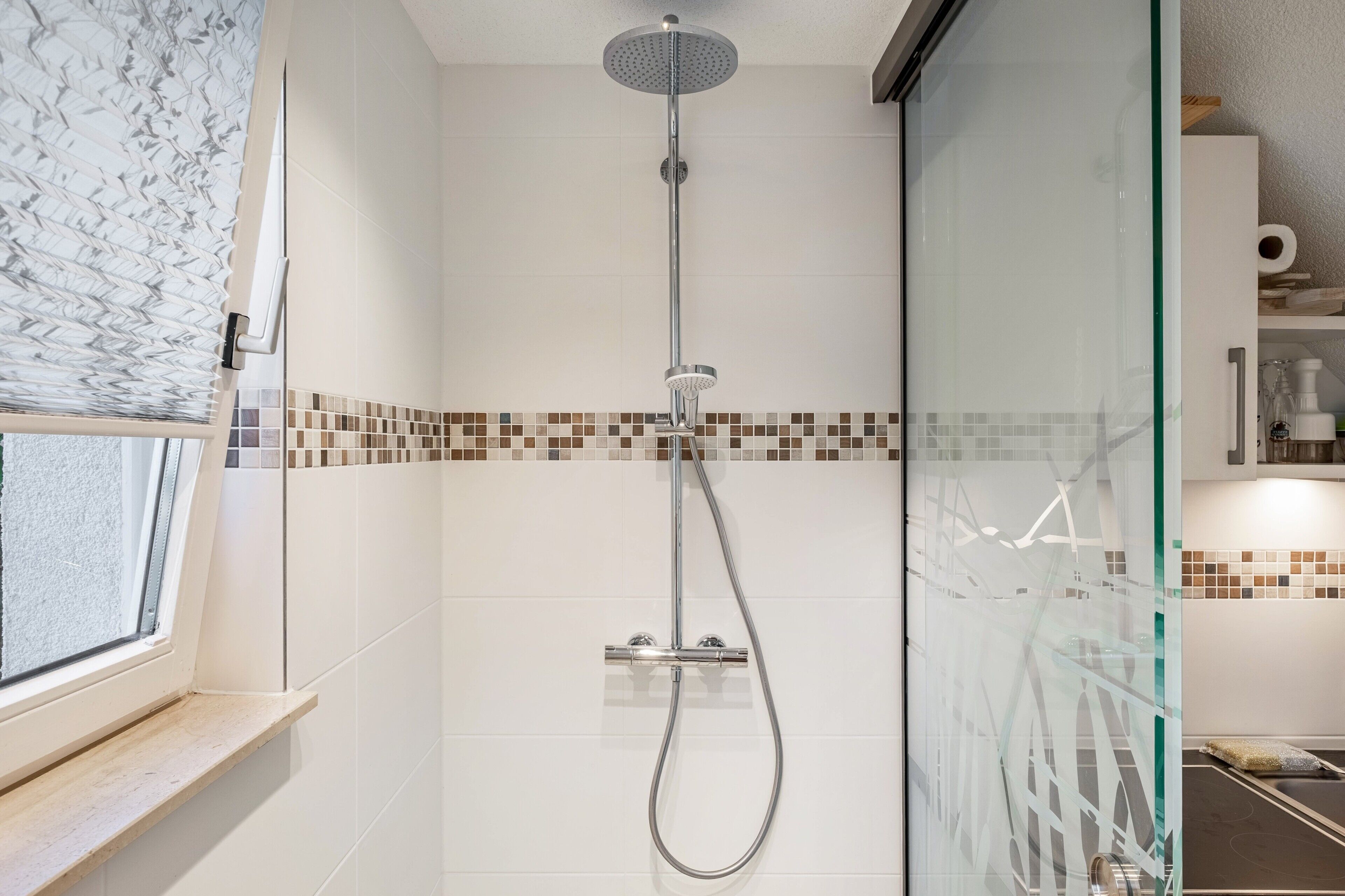Ducha con mampara de vidrio, ventana con vistas y pared de azulejos con detalle de mosaico.