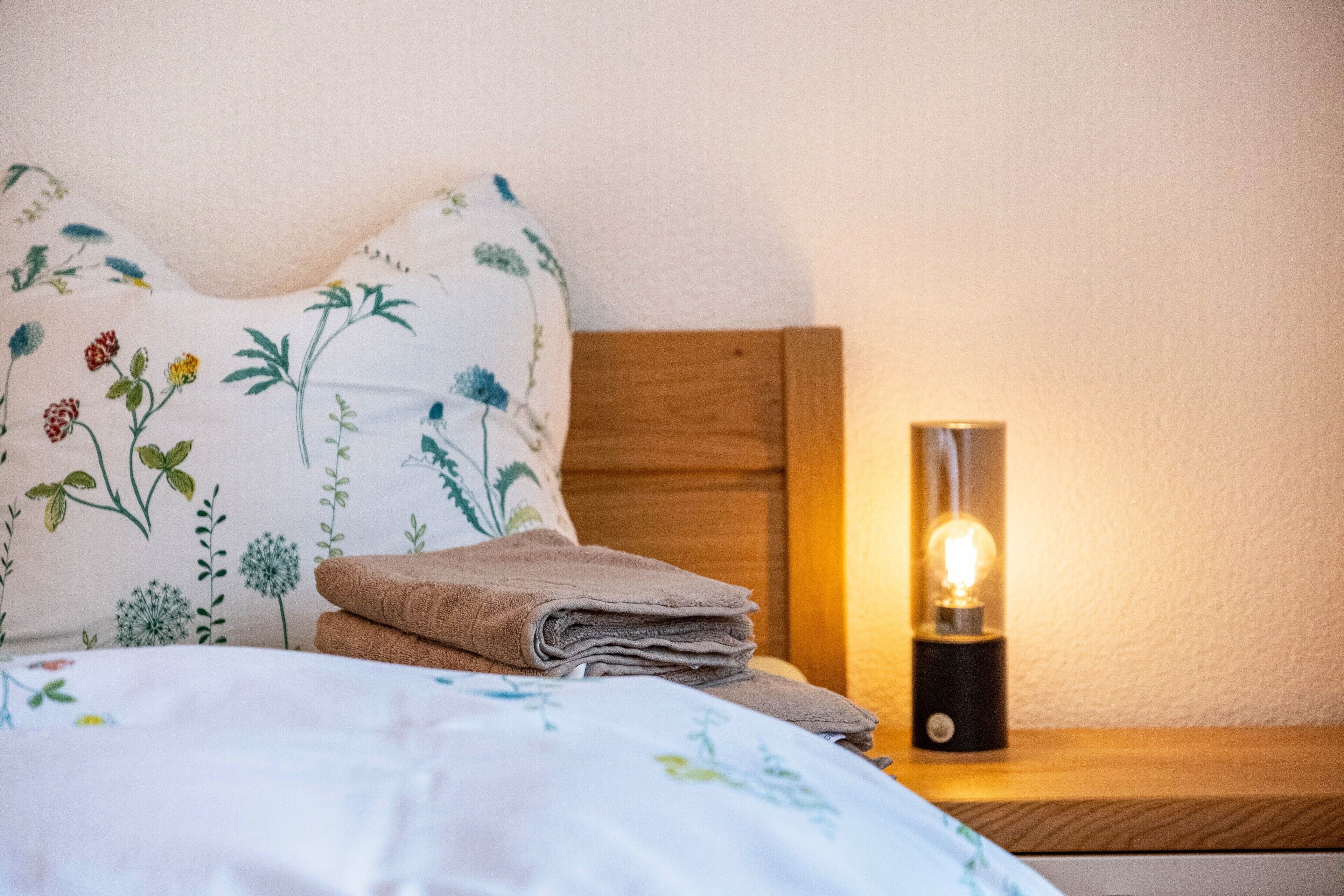 A neatly made bed with a floral pillow, a stack of folded towels, and a bedside table with a lamp.