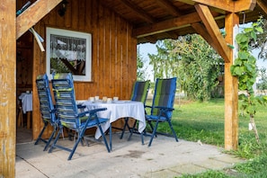 Eine hölzerne Außenkonstruktion mit Tisch und Stühlen, ein Fenster mit Vorhang sowie Blick auf Grünfläche.
