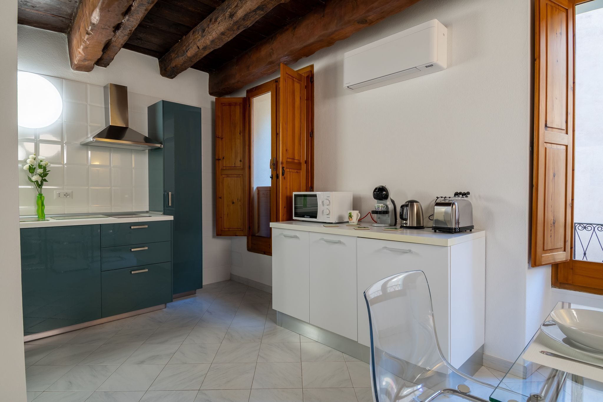 A modern kitchen with a wooden ceiling, white cabinets, a microwave, a kettle, and a glass chair.