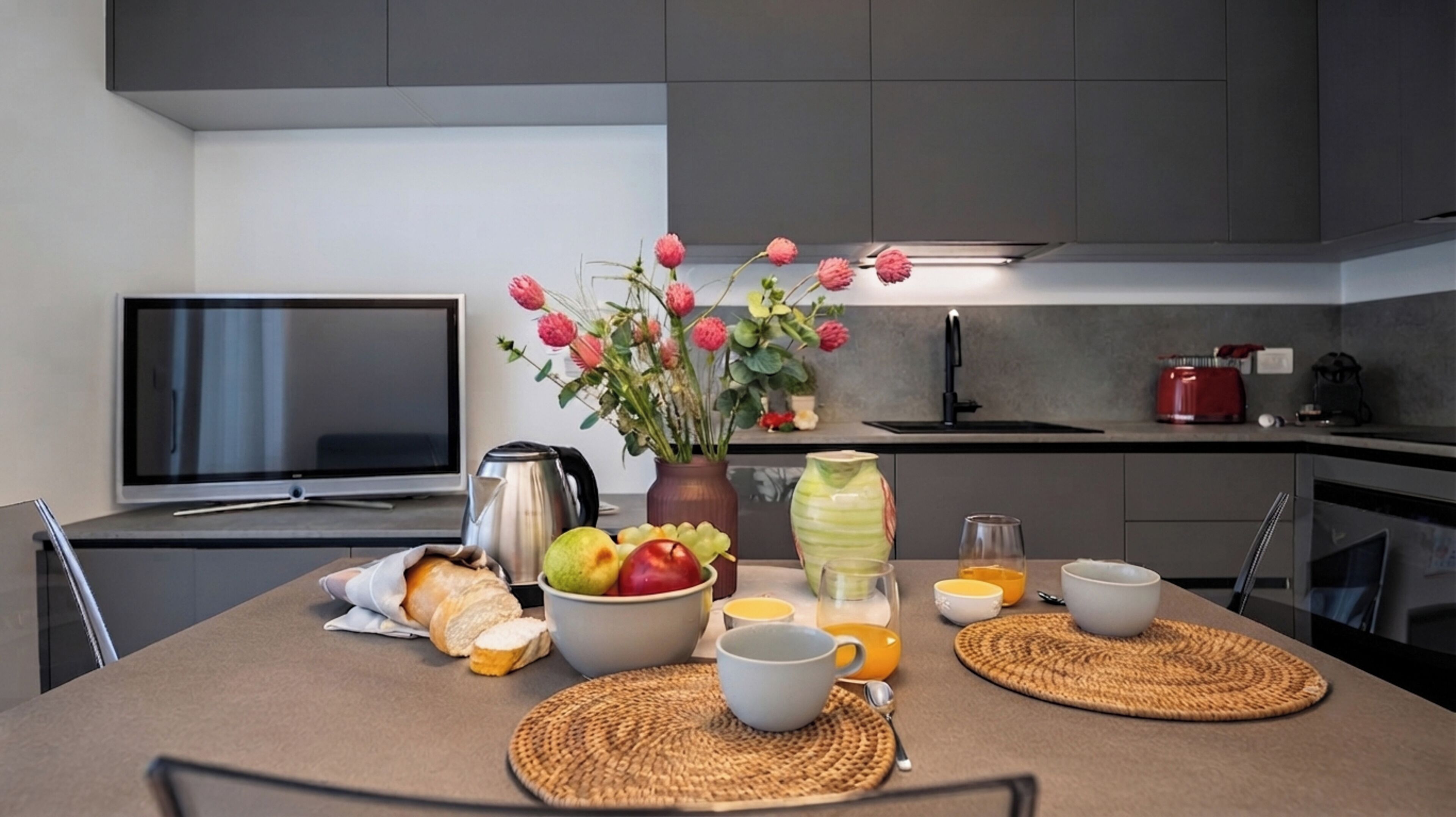 A modern kitchen with a flat-screen TV, a coffee maker, a fruit bowl, a vase with flowers, and a red kettle.