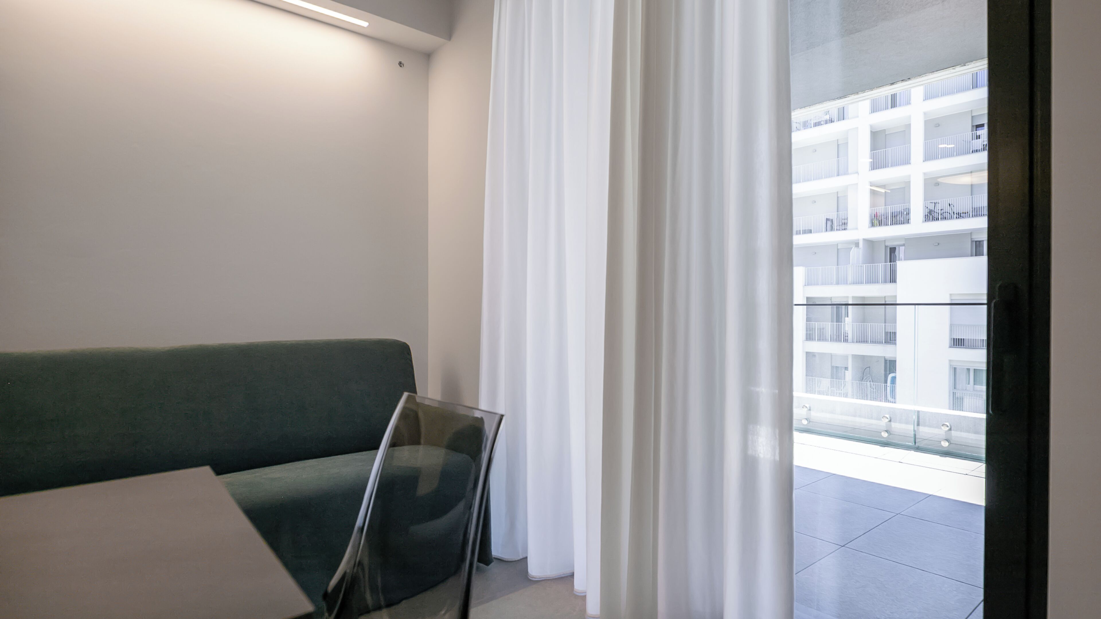 Ein modernes Zimmer mit einem grünen Sofa, einem Glastisch und Blick auf ein weißes Gebäude durch transparente Vorhänge.