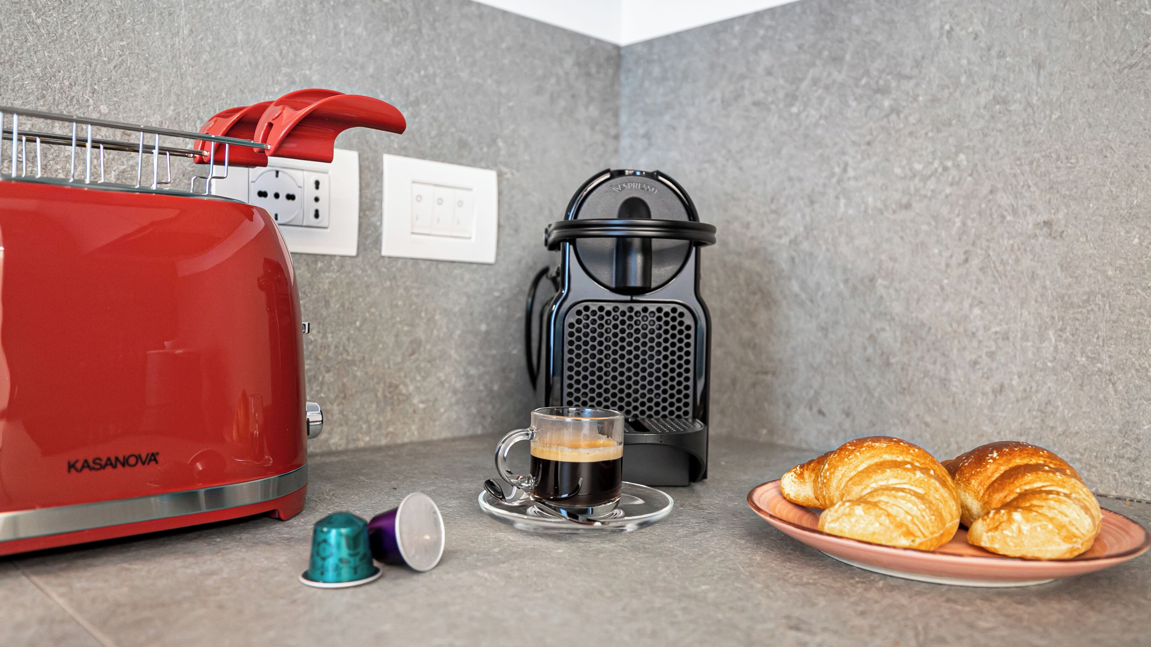 A red coffee maker, a black espresso machine, a glass cup with coffee, and a plate with croissants on a kitchen counter.
