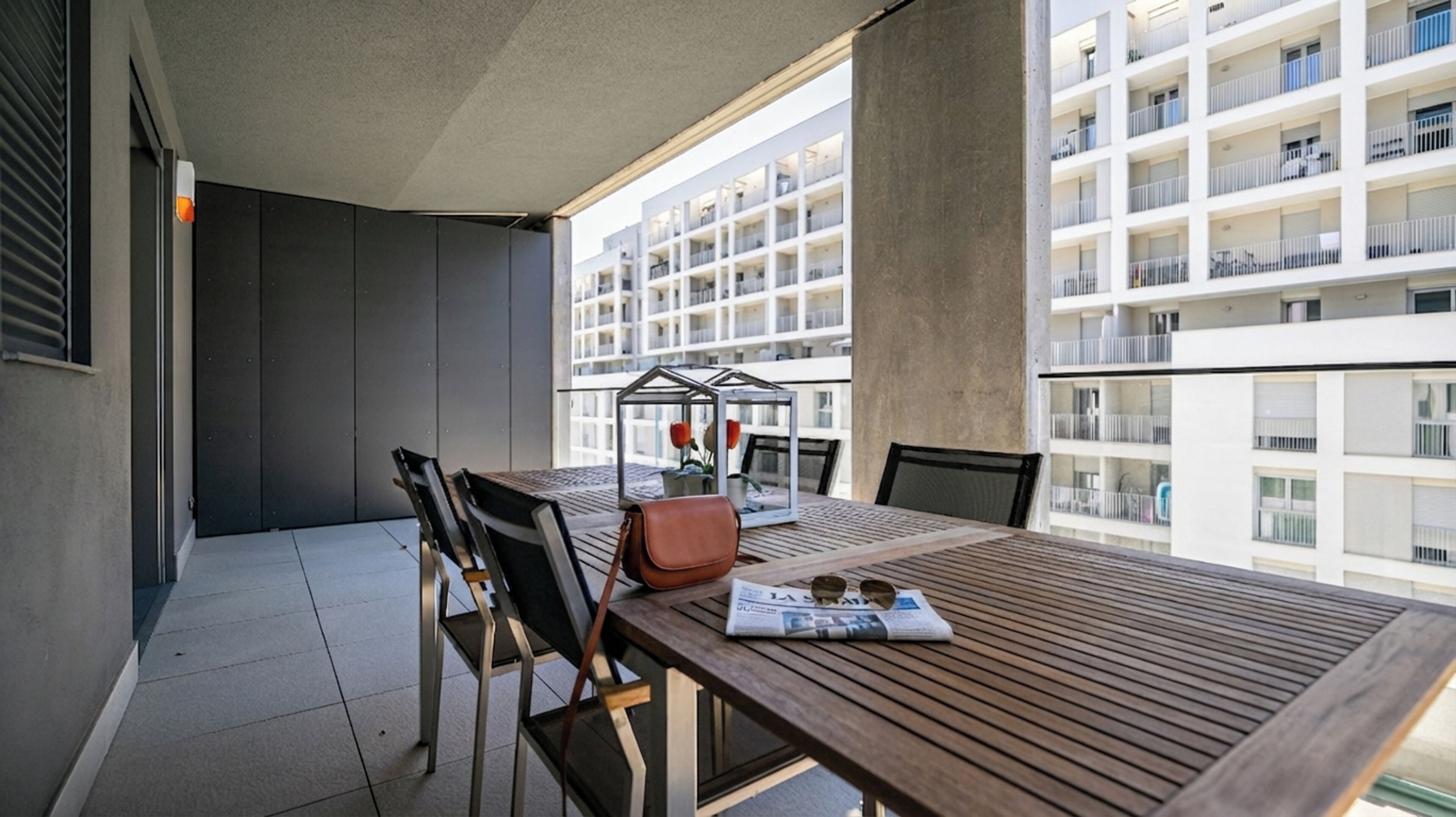 Un balcon avec une table en bois, des chaises, une structure en verre abritant des fleurs et une vue sur un immeuble à plusieurs étages.