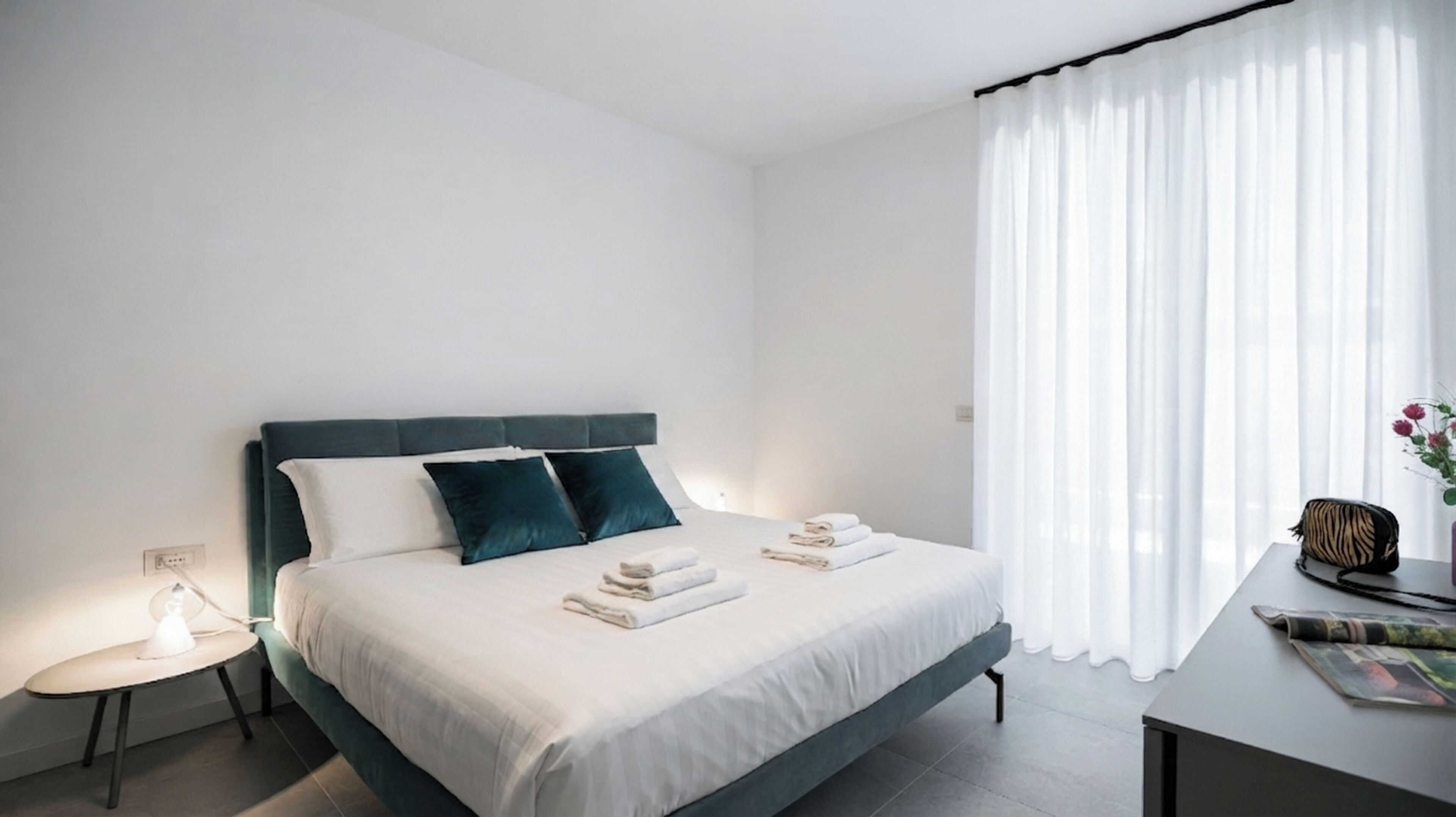 Una habitación de hotel moderna con una cama, una mesita de noche, una lámpara y un ventanal con cortinas.